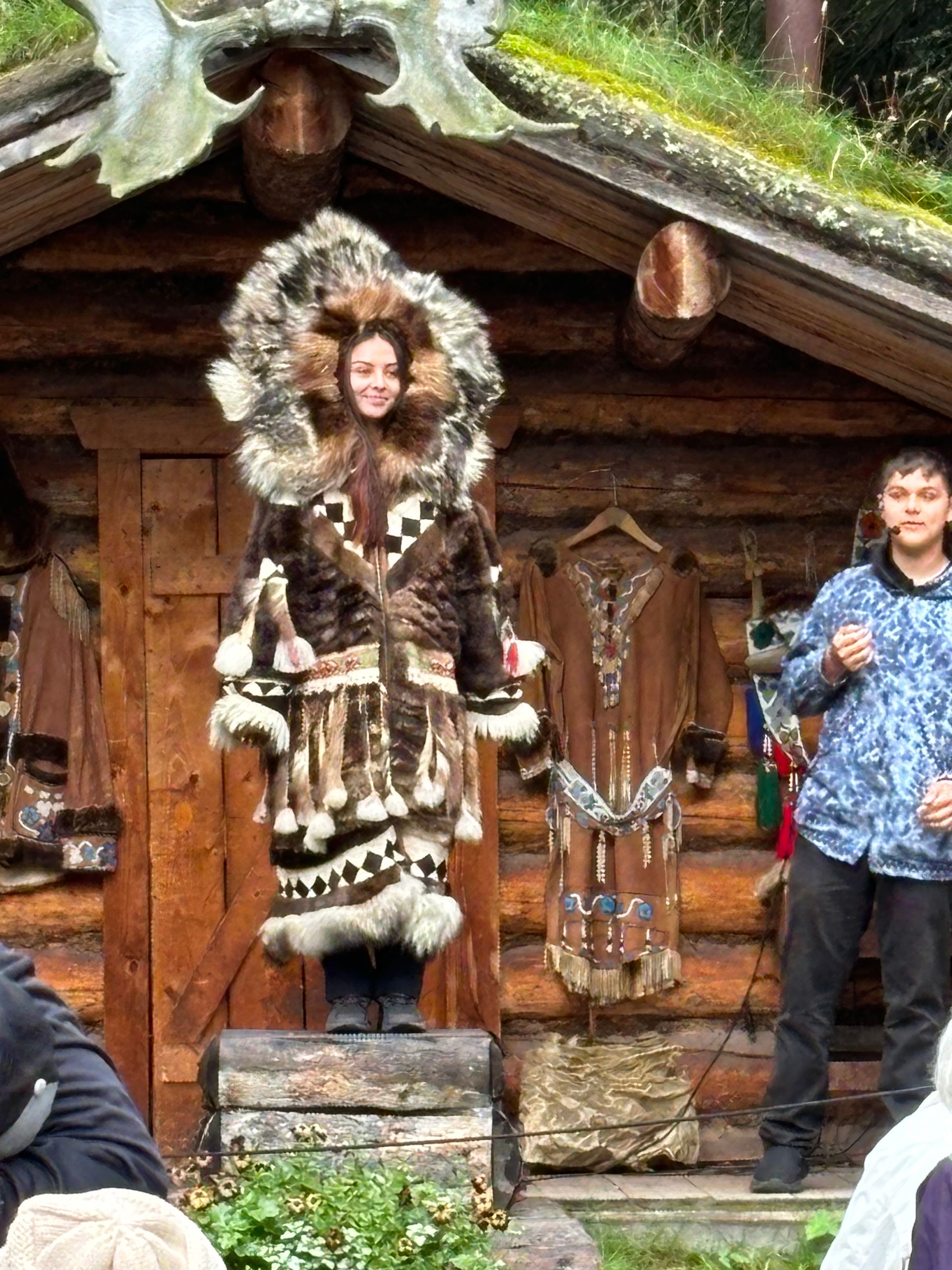 A person in traditional fur clothing stands on a wooden platform in front of a log building, beside a standing person.