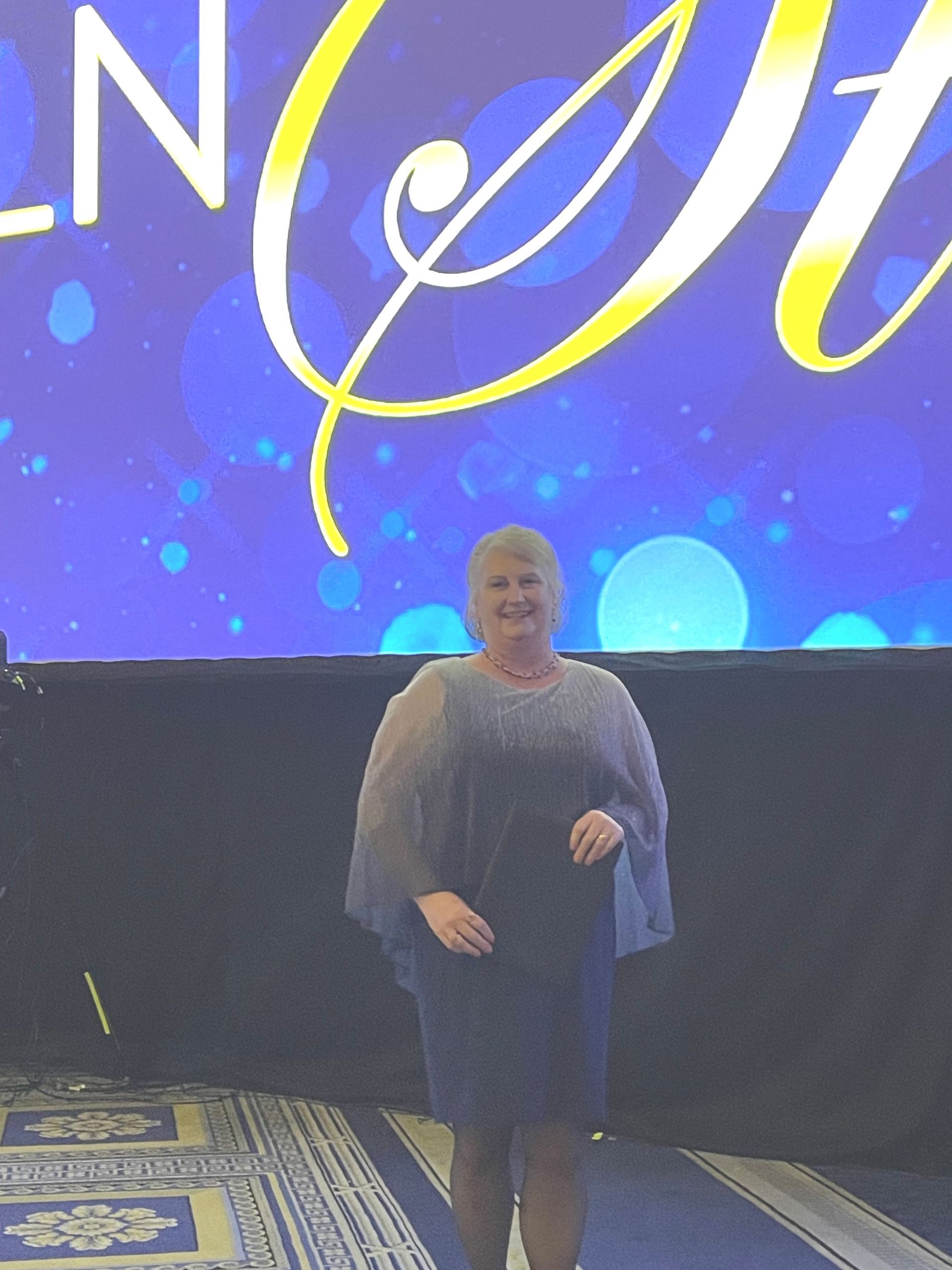 A person smiling while standing on a stage in front of a blue background with a large yellow sign.