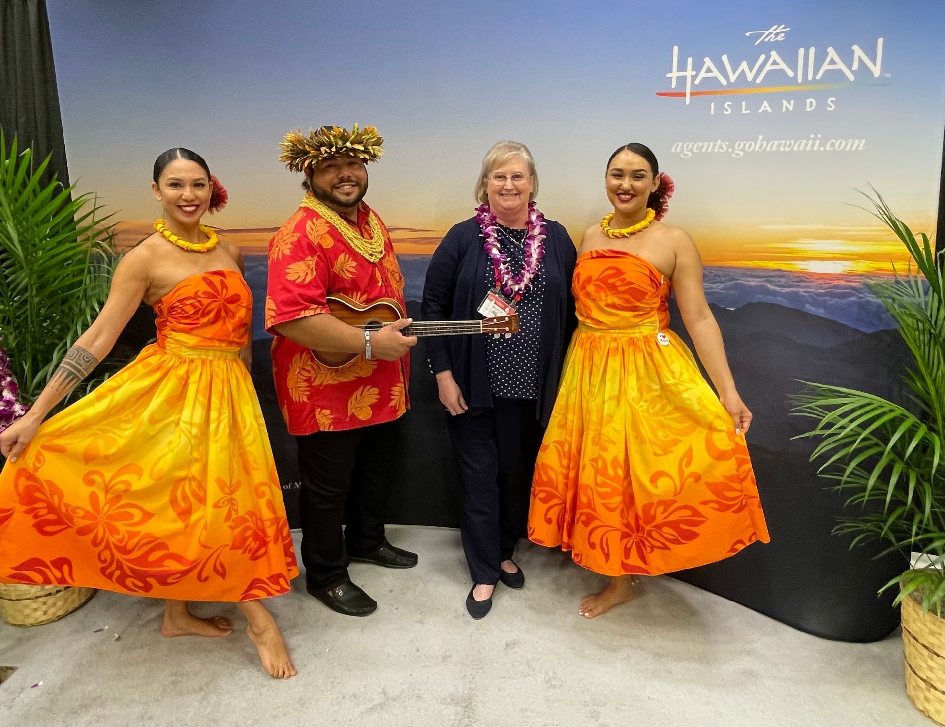 Four people posing in front of a Hawaiian Islands banner, including two dancers in yellow-orange skirts and a musician.