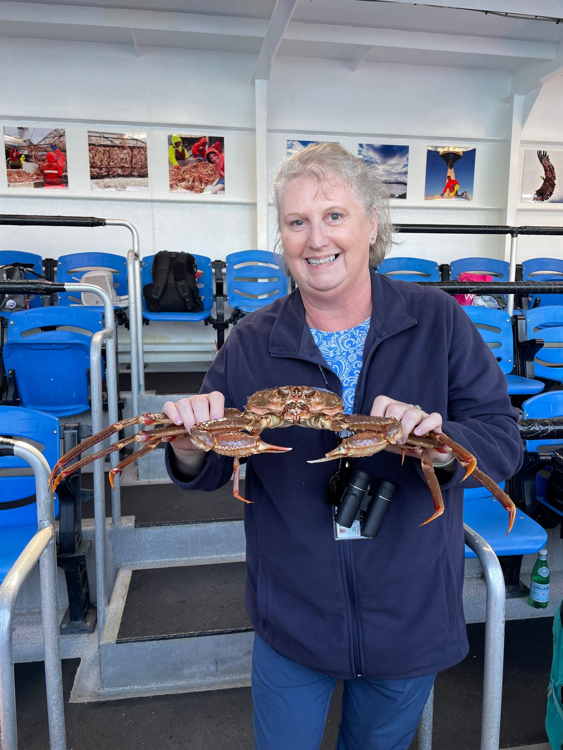 A smiling person in a blue fleece jacket holds a large crab in front of rows of blue seats on a boat.
