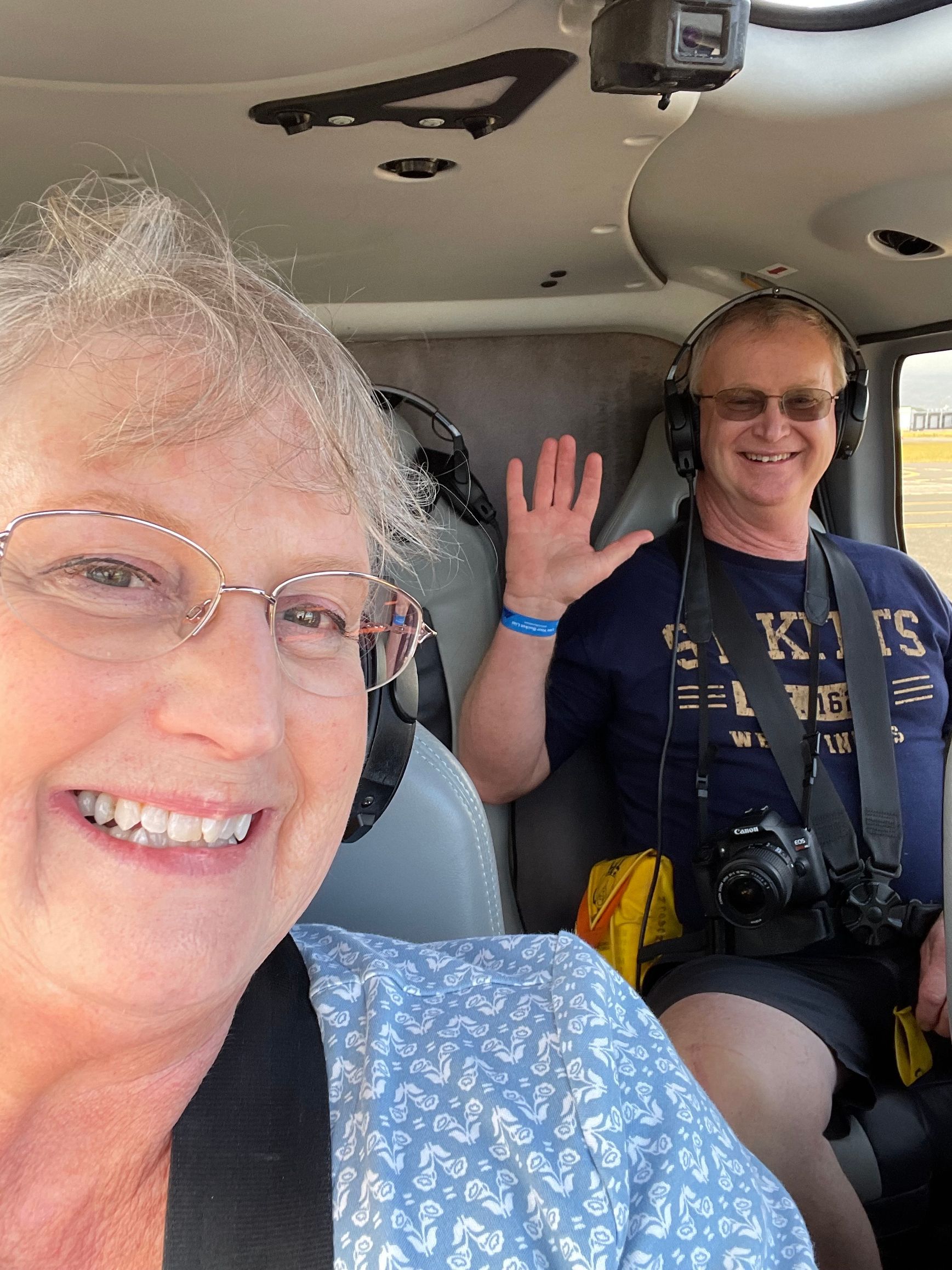 A woman in a blue patterned top smiles for a selfie while a man wearing headphones and a camera waves from behind her.