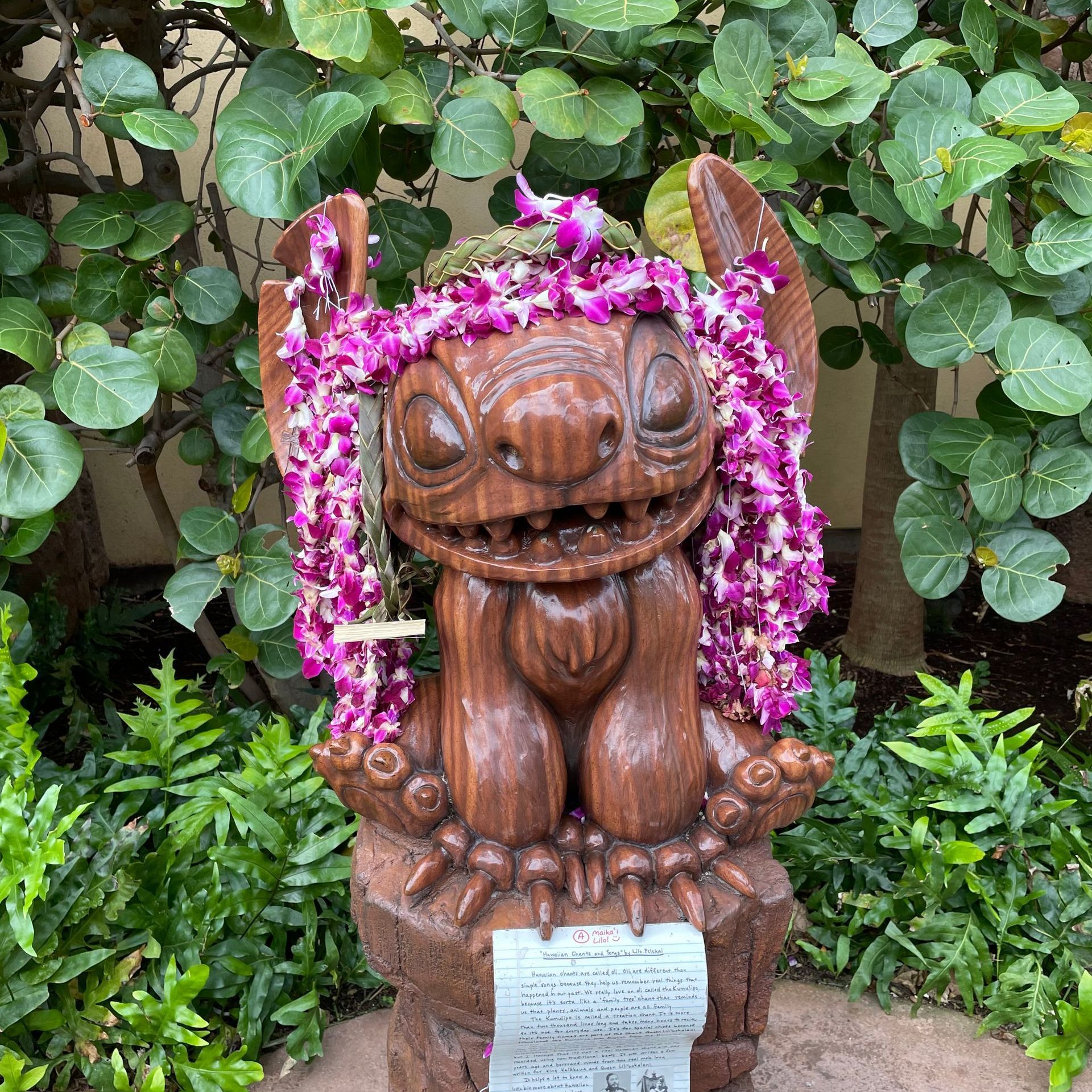 A wooden statue of Stitch decorated with purple flower leis, standing on a stone base among lush green tropical plants.