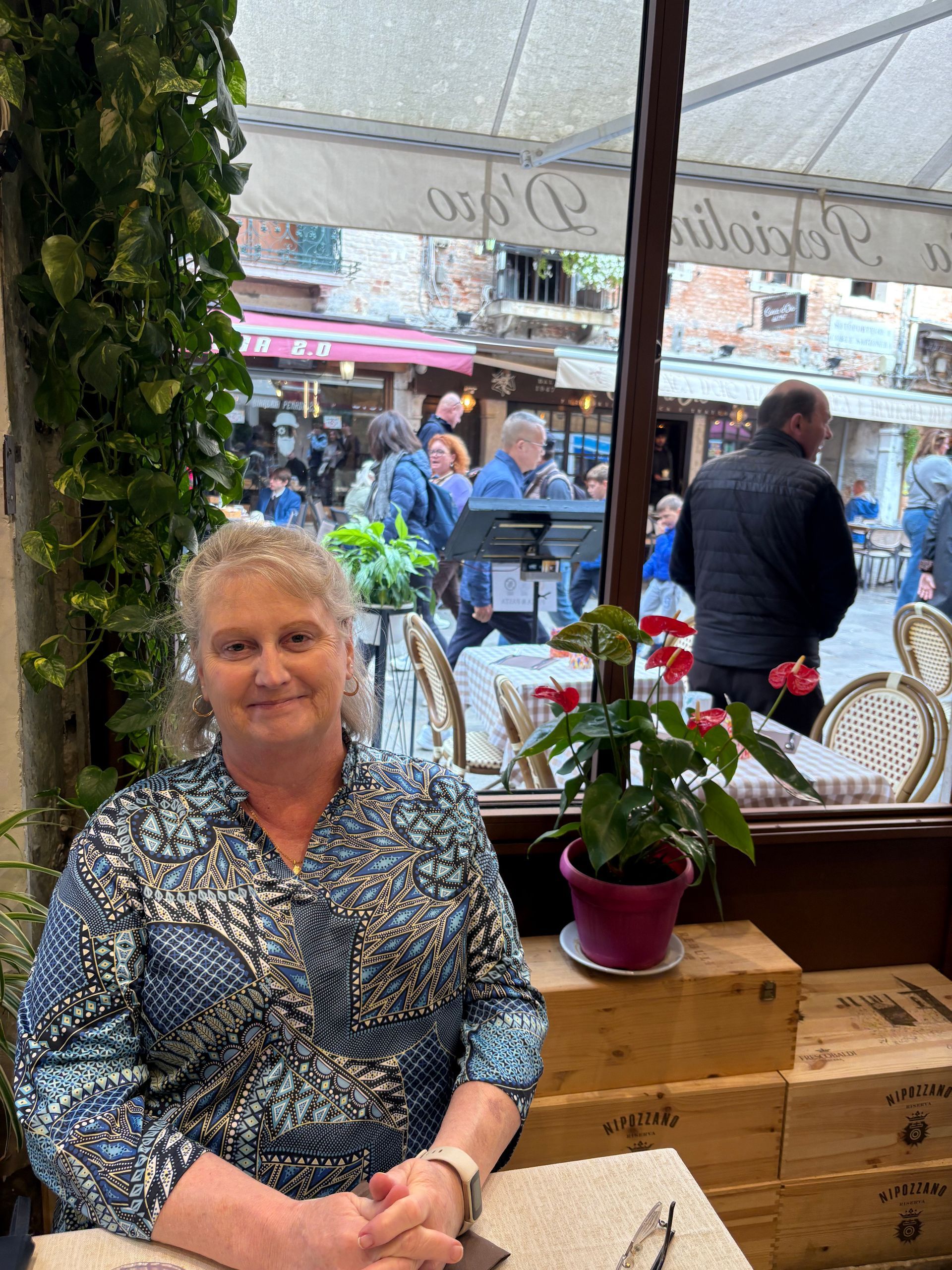 A person sits at a table in an outdoor cafe in a cobblestone street, smiling with their hands clasped.