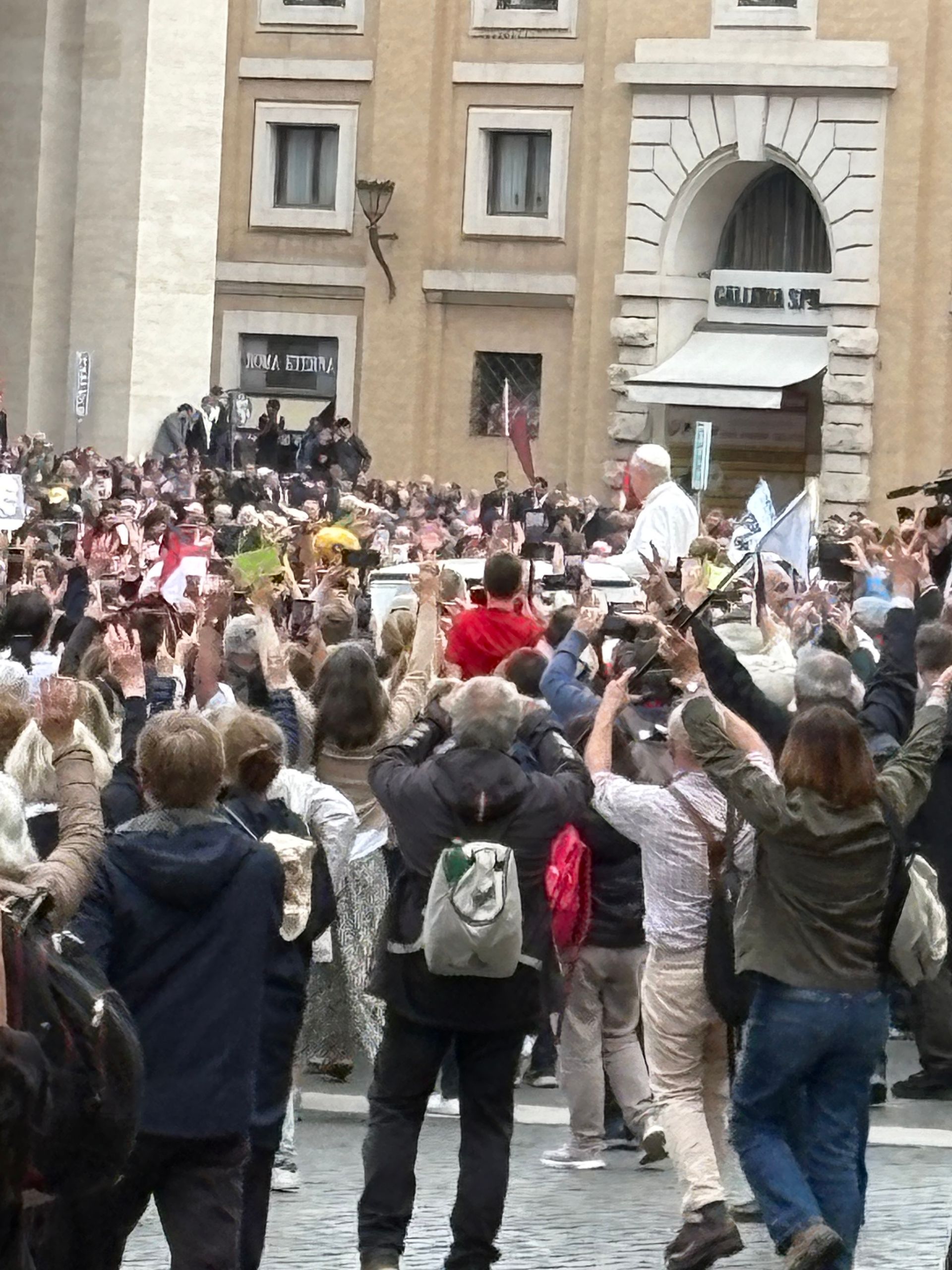 Pope Francis waving from a vehicle to a cheering, flag-waving crowd gathered in a city square.
