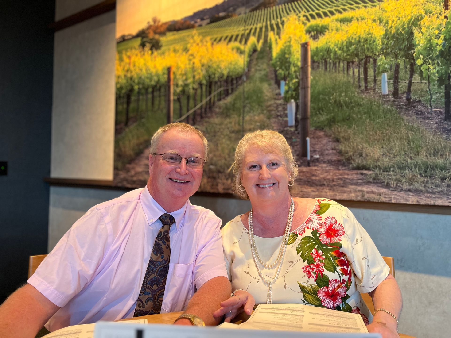 A smiling couple sits at a table in a room with a large wall mural of a vineyard.