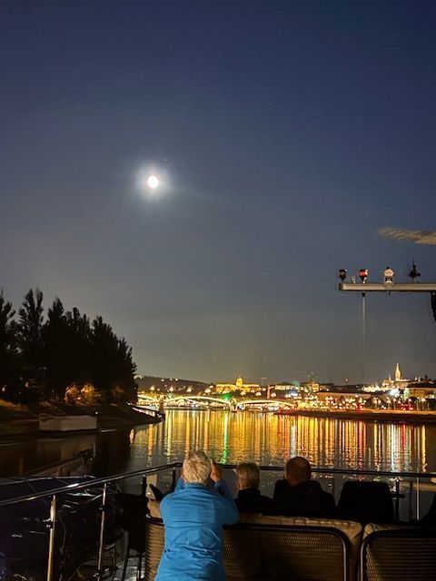 Three people on a boat at night watch the bright moon shining over a brightly lit city skyline reflecting on the water.