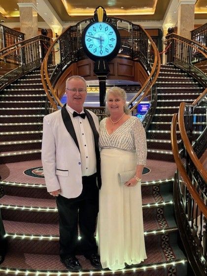 A couple in formal attire stands smiling on a grand staircase in a luxurious cruise ship atrium with a central clock.