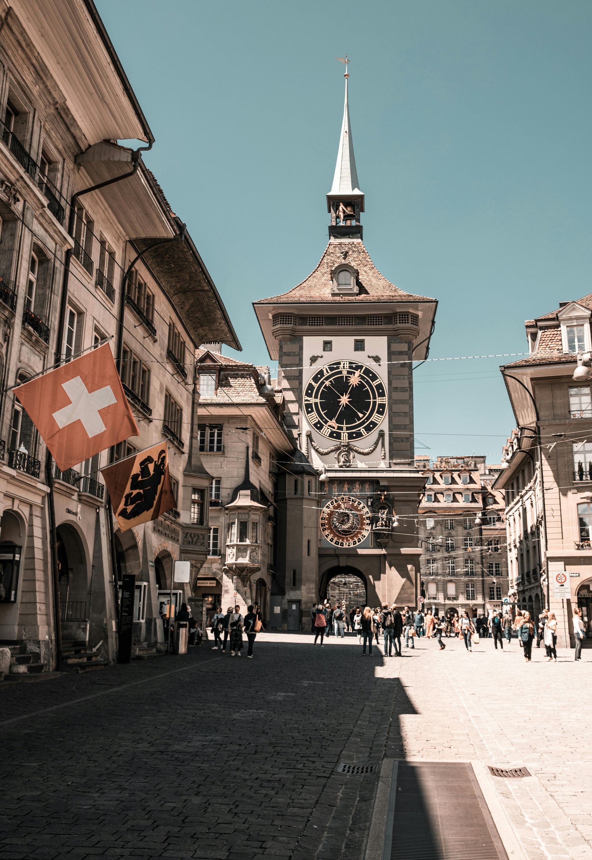 A clock tower in a city with a swiss flag flying in front of it.