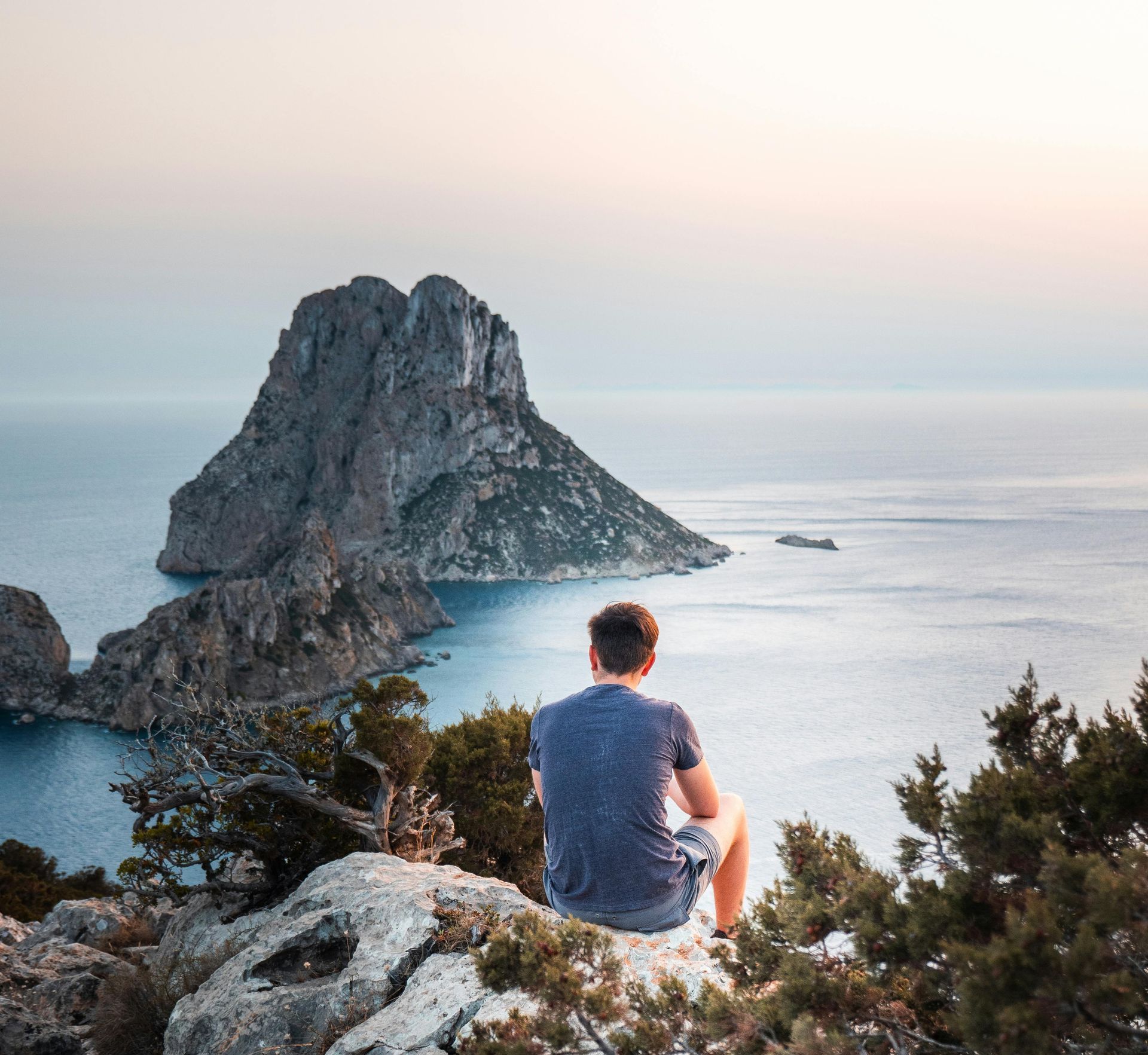 A person sits on a rocky cliff overlooking Es Vedrà, a distinctive limestone island in the Mediterranean Sea at sunset.