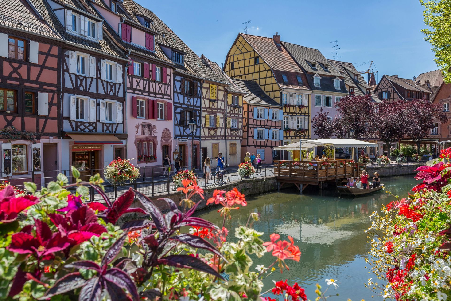 Colorful half-timbered houses line a canal in Colmar, France, with vibrant flower boxes and a small boat in the water.