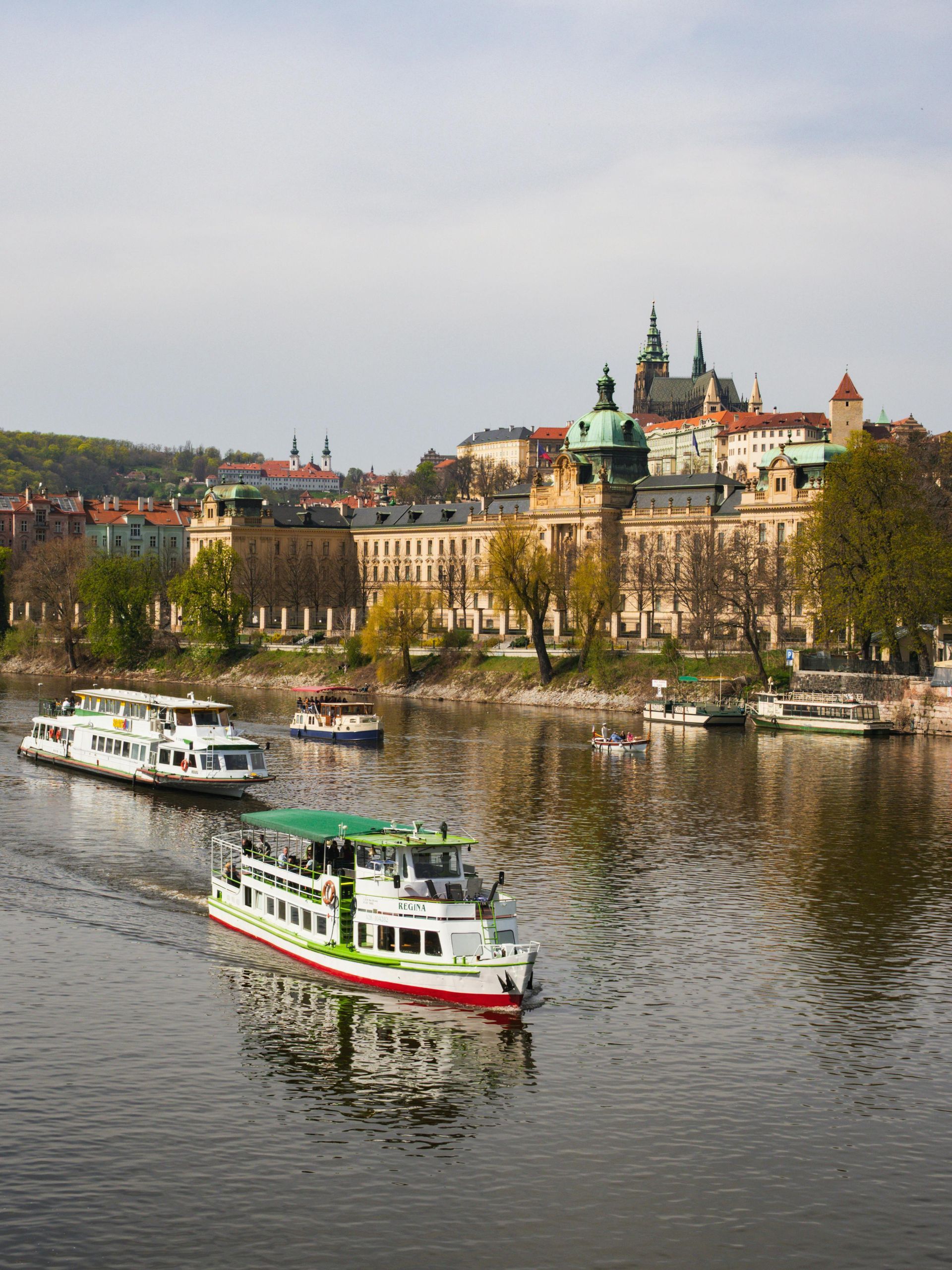 Two tour boats travel on the Vltava River in Prague with the Strakonice Academy and Prague Castle in the background.