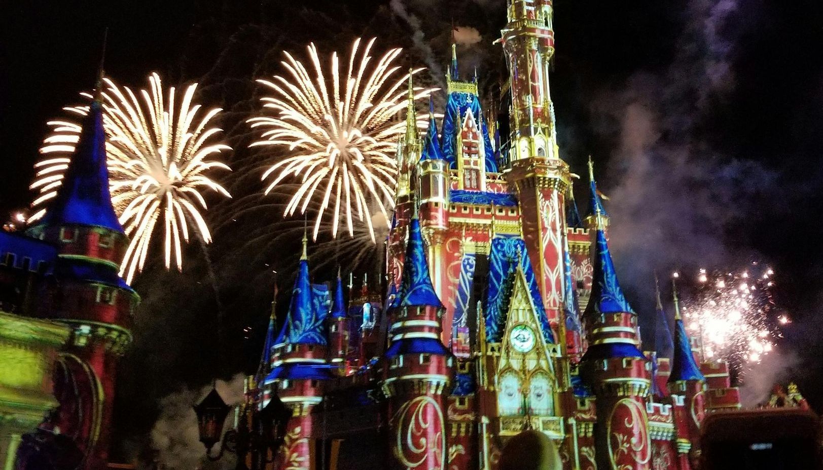 Fireworks explode behind the brightly lit Cinderella Castle at night at a Disney theme park.