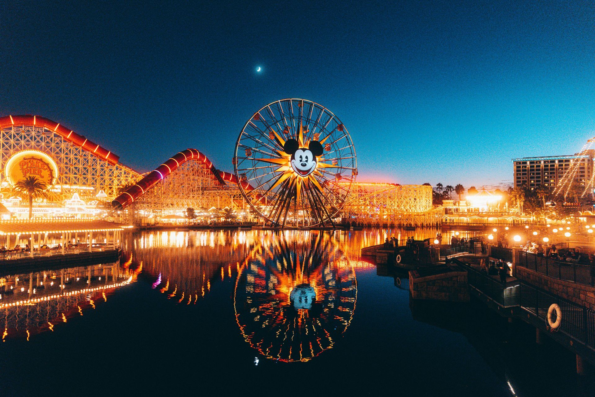 A mickey mouse ferris wheel is reflected in the water at an amusement park.