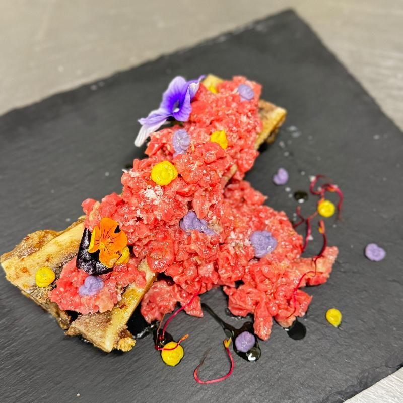 Piatto di tartare di manzo su una lavagna nera, guarnito con fiori e salse.