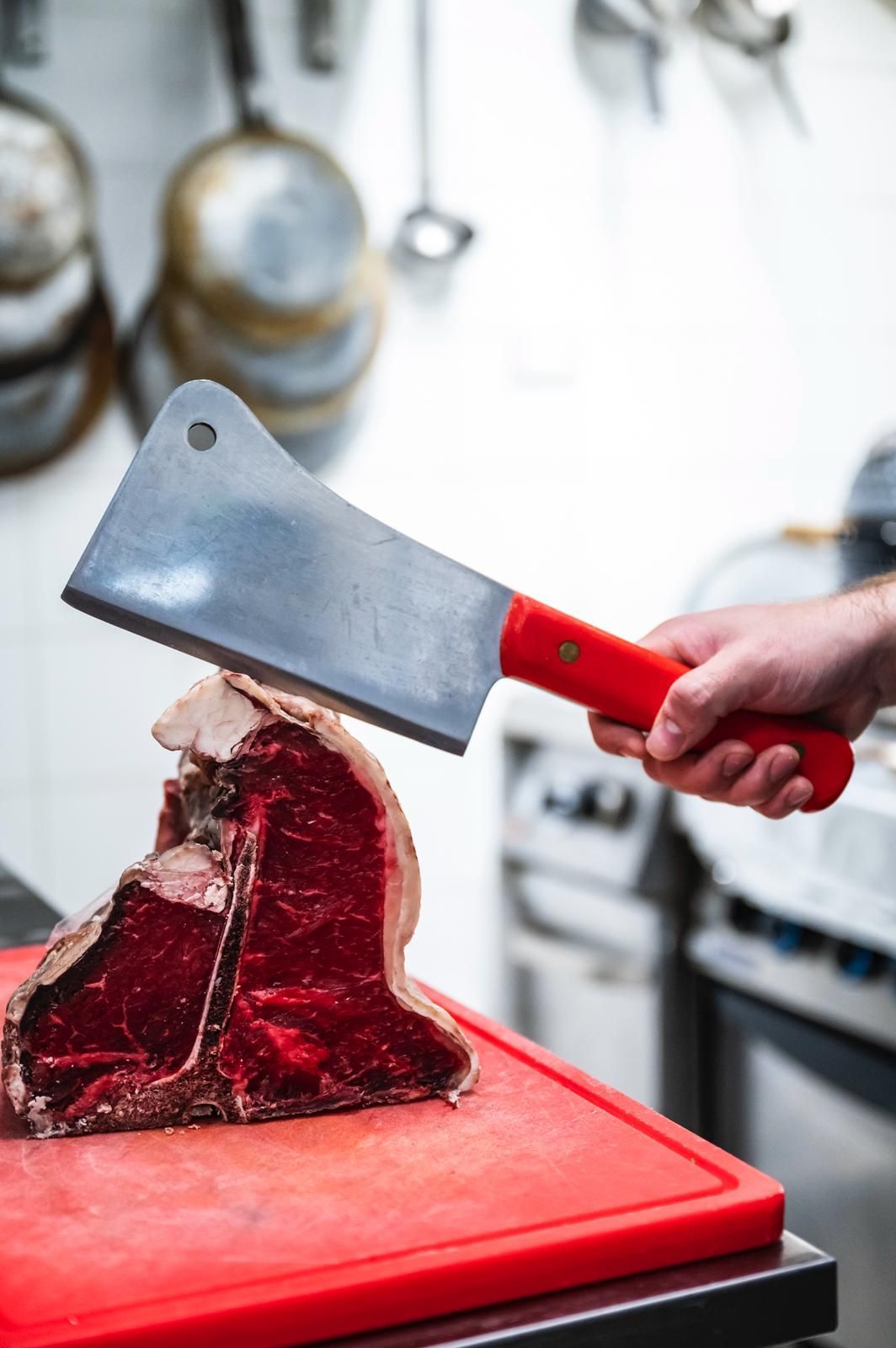 Mano che regge una mannaia sopra una bistecca T-bone cruda su un tagliere rosso in cucina.