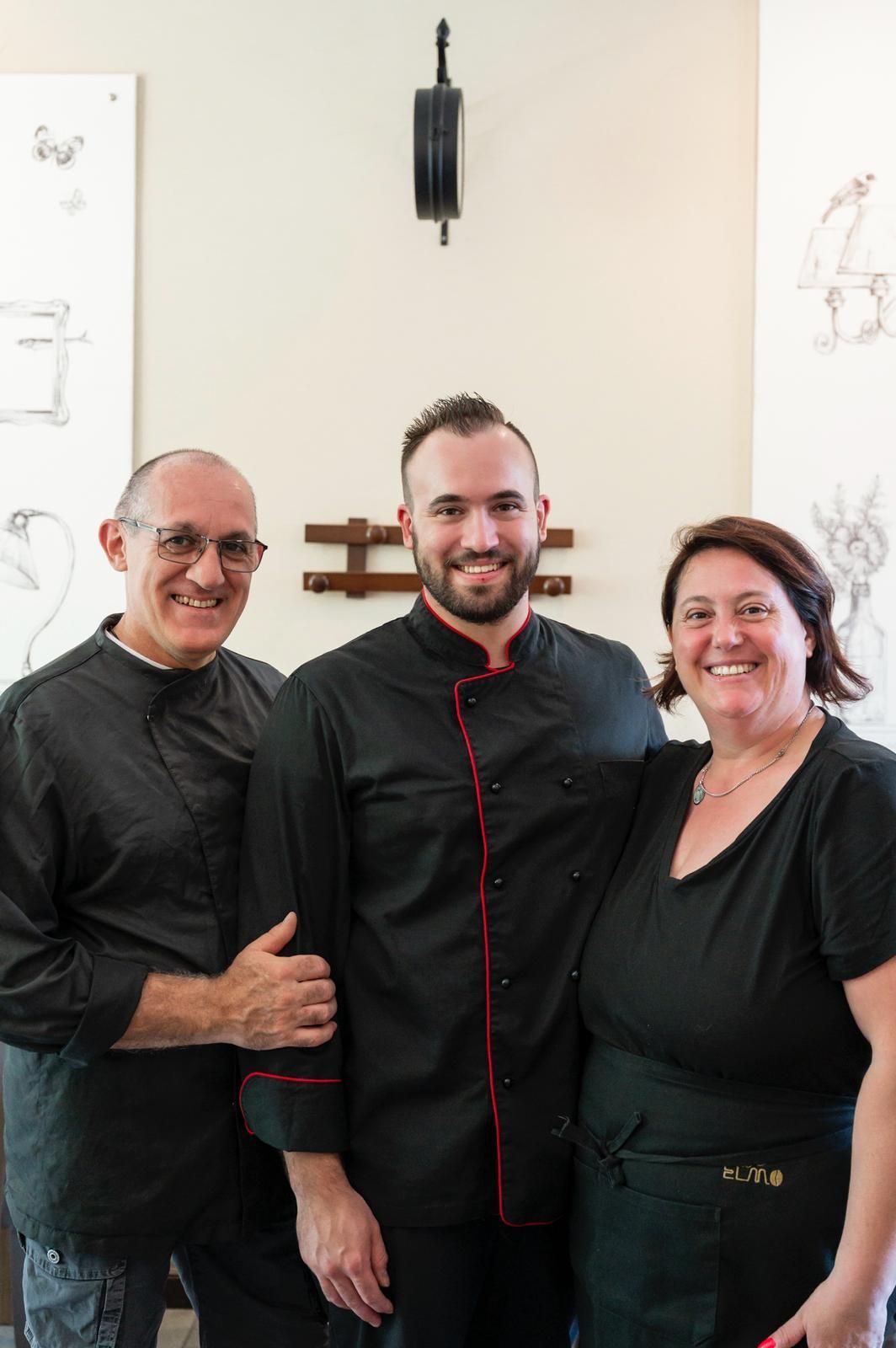 Tre persone vestite di nero sono in piedi in un ristorante; l'uomo al centro sorride.