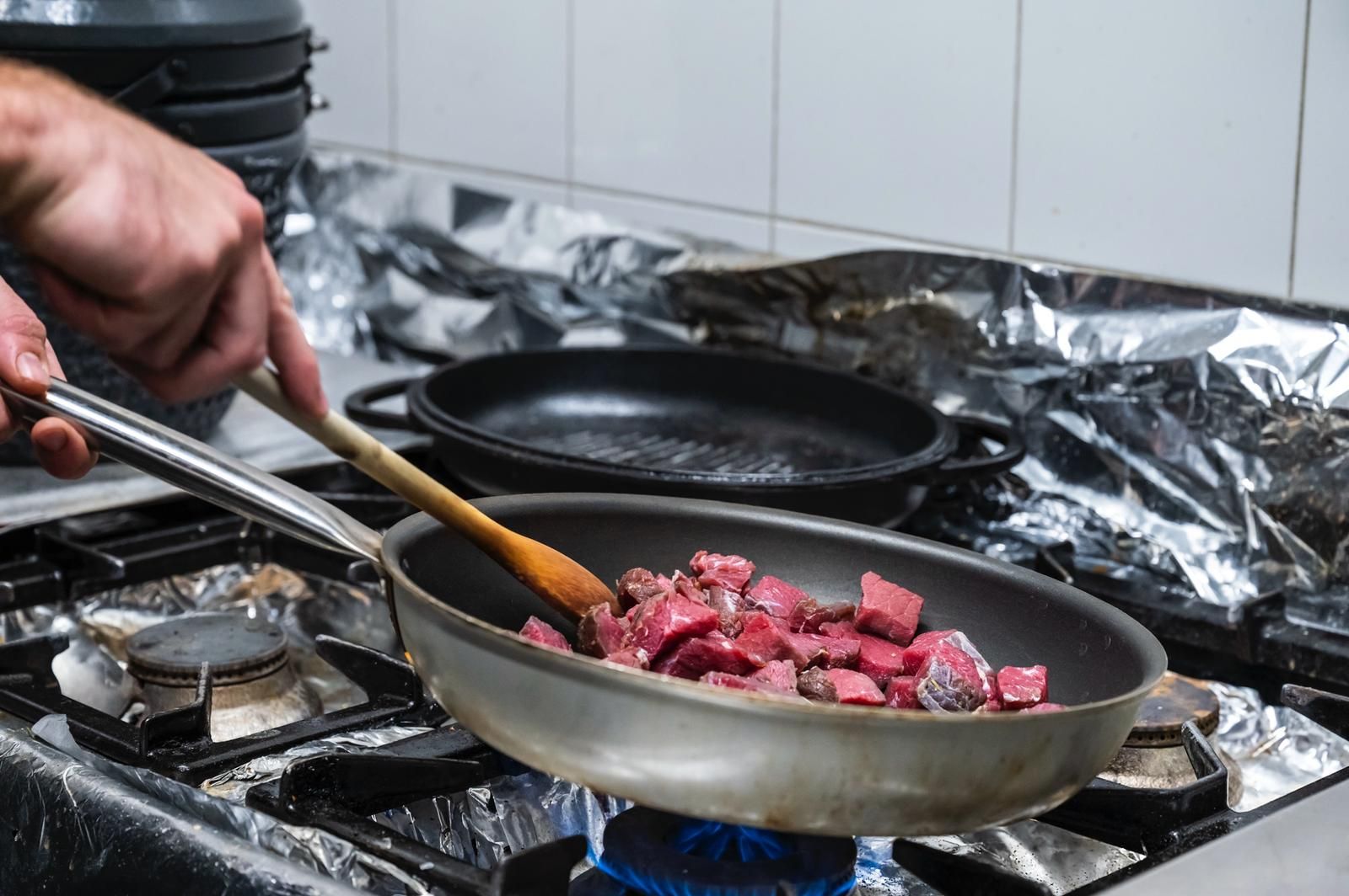 Persona che mescola cubetti di carne cruda in una padella su un fornello a gas, in cucina.