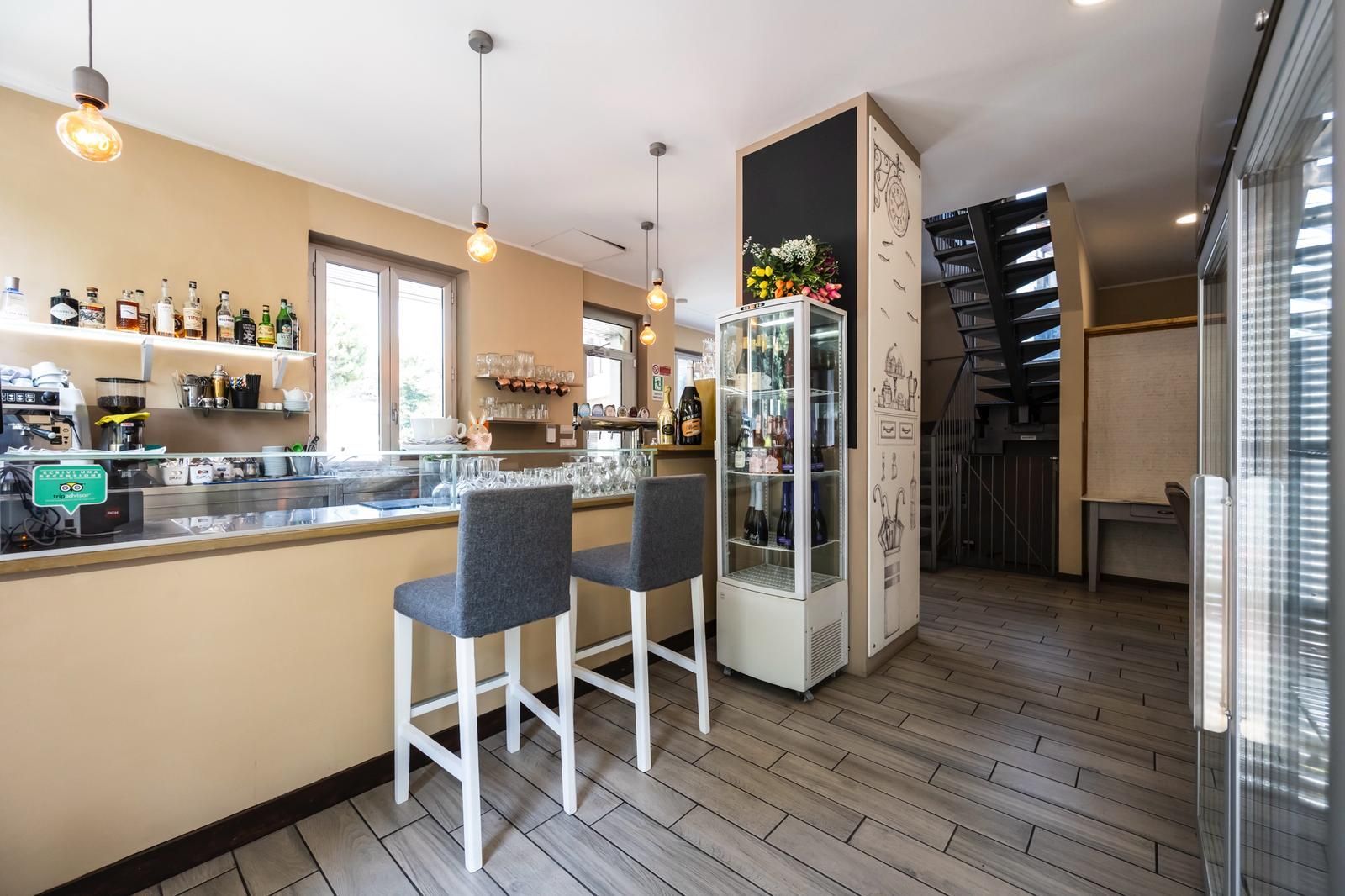 Vista interna di un bar con sgabelli, bancone, bottiglie e una scala sullo sfondo.