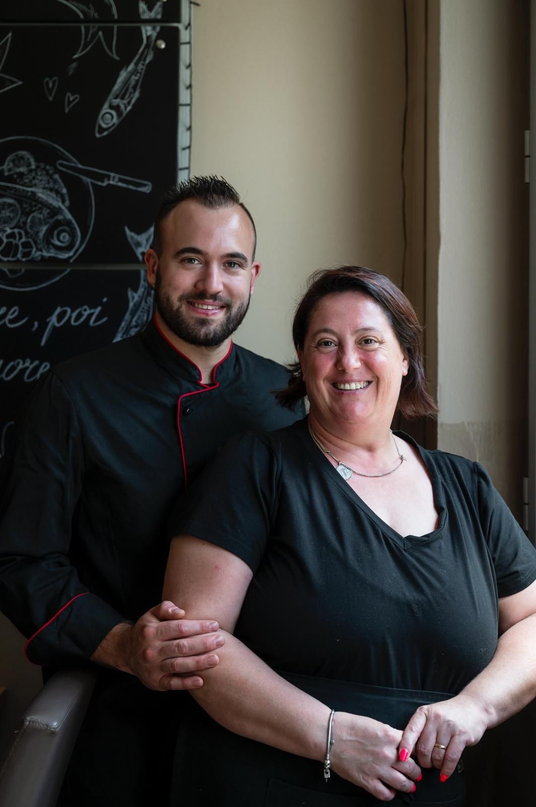Due ristoratori sorridenti: un uomo con la giacca da chef e una donna, in piedi vicino a una lavagna.