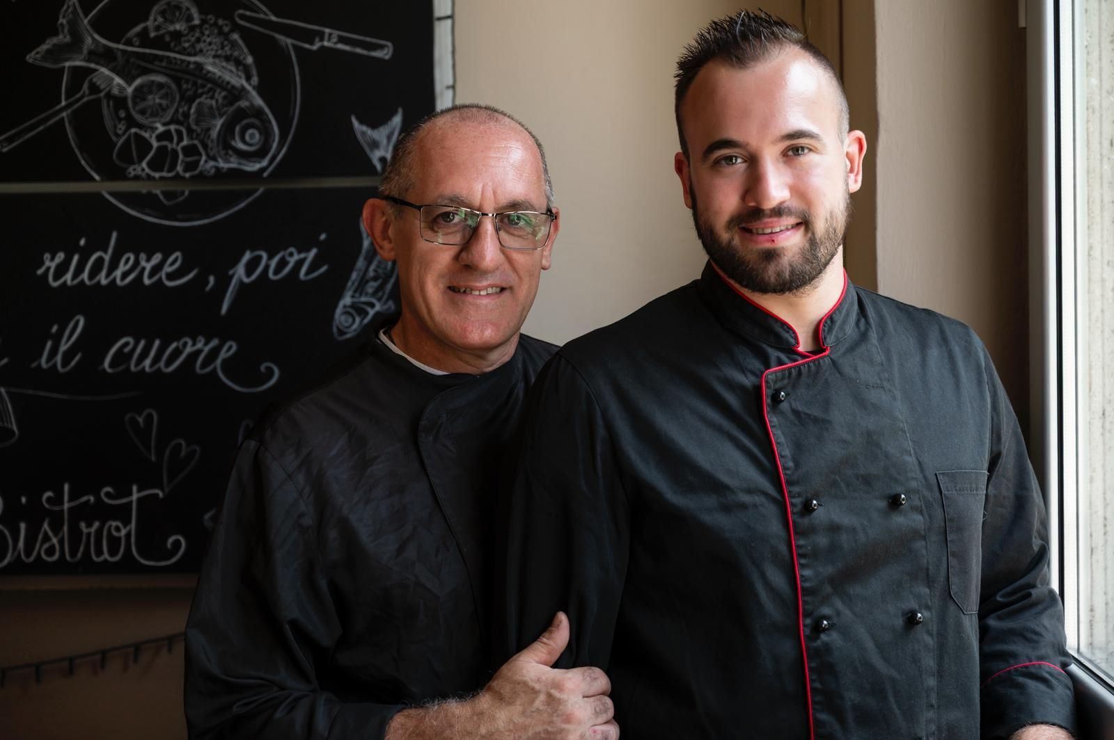 Due uomini in camice da cuoco nero, posano insieme in un interno; uno più anziano, l'altro più giovane.