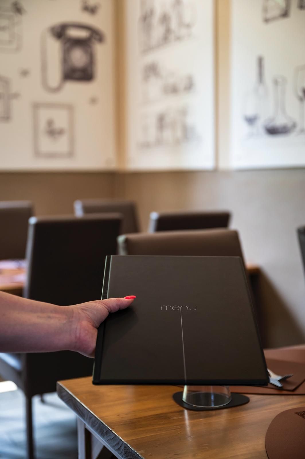 Mano che regge un menù nero su un tavolo di legno in un ristorante, decorato con opere d'arte alle pareti.