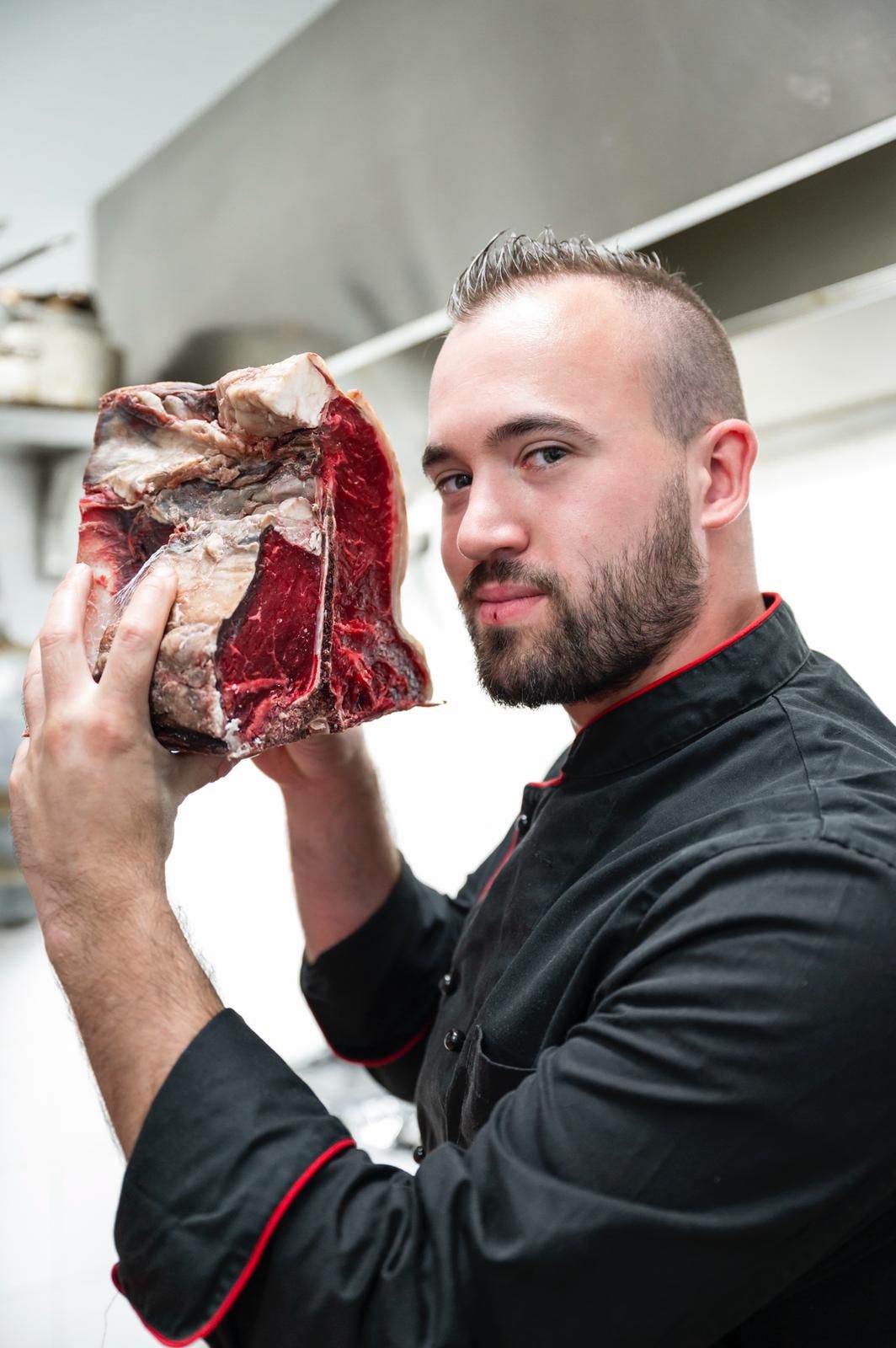 Uno chef tiene in mano una grande bistecca frollata a secco in cucina. Guarda la telecamera con un sorriso.