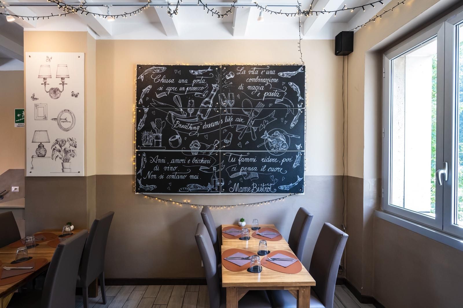 Interno di un ristorante con menù scritto sulla lavagna, tavoli apparecchiati per mangiare e una finestra.