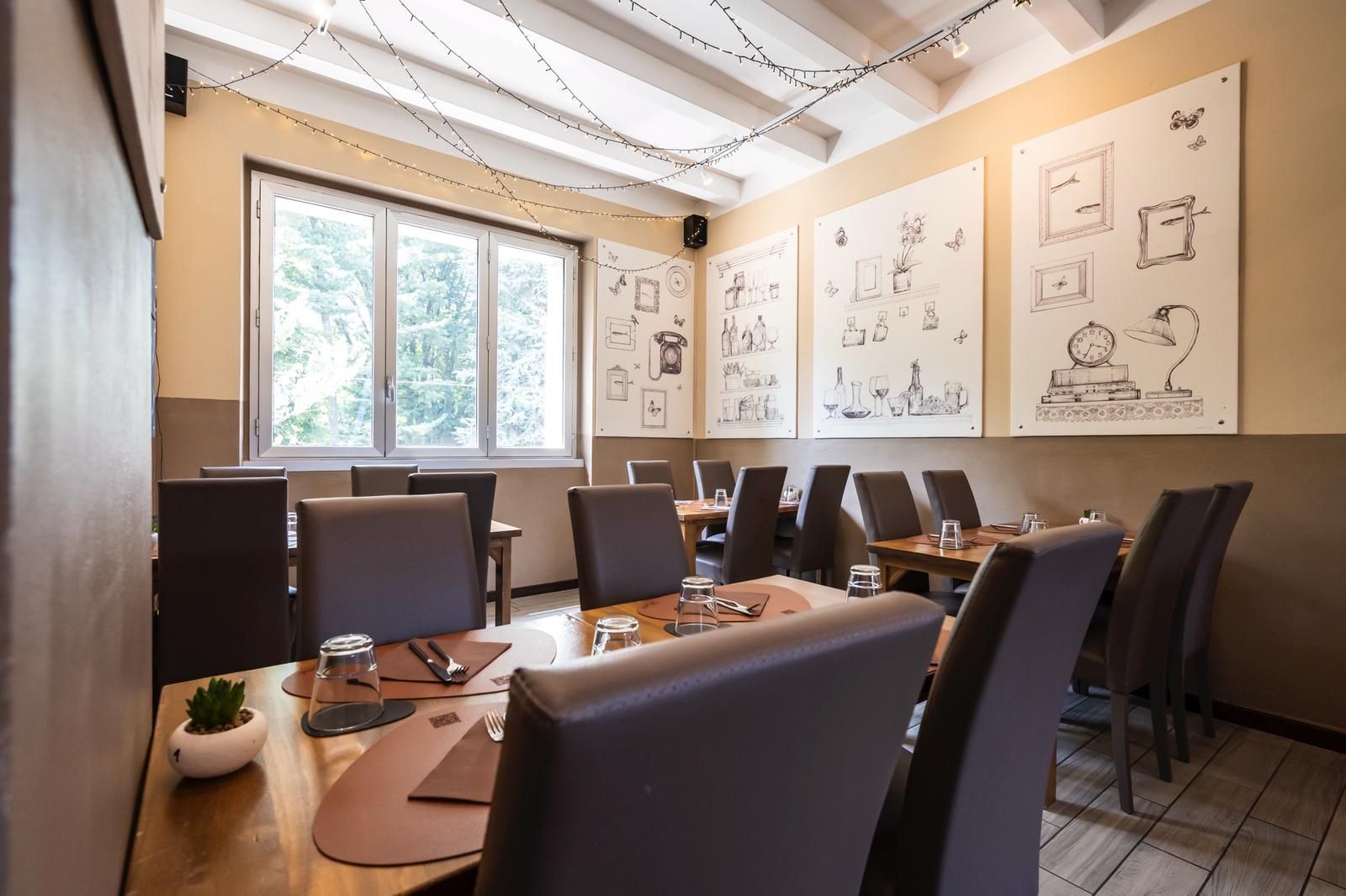 Sala da pranzo del ristorante con tavoli apparecchiati, sedie marroni, disegni alle pareti e una finestra.