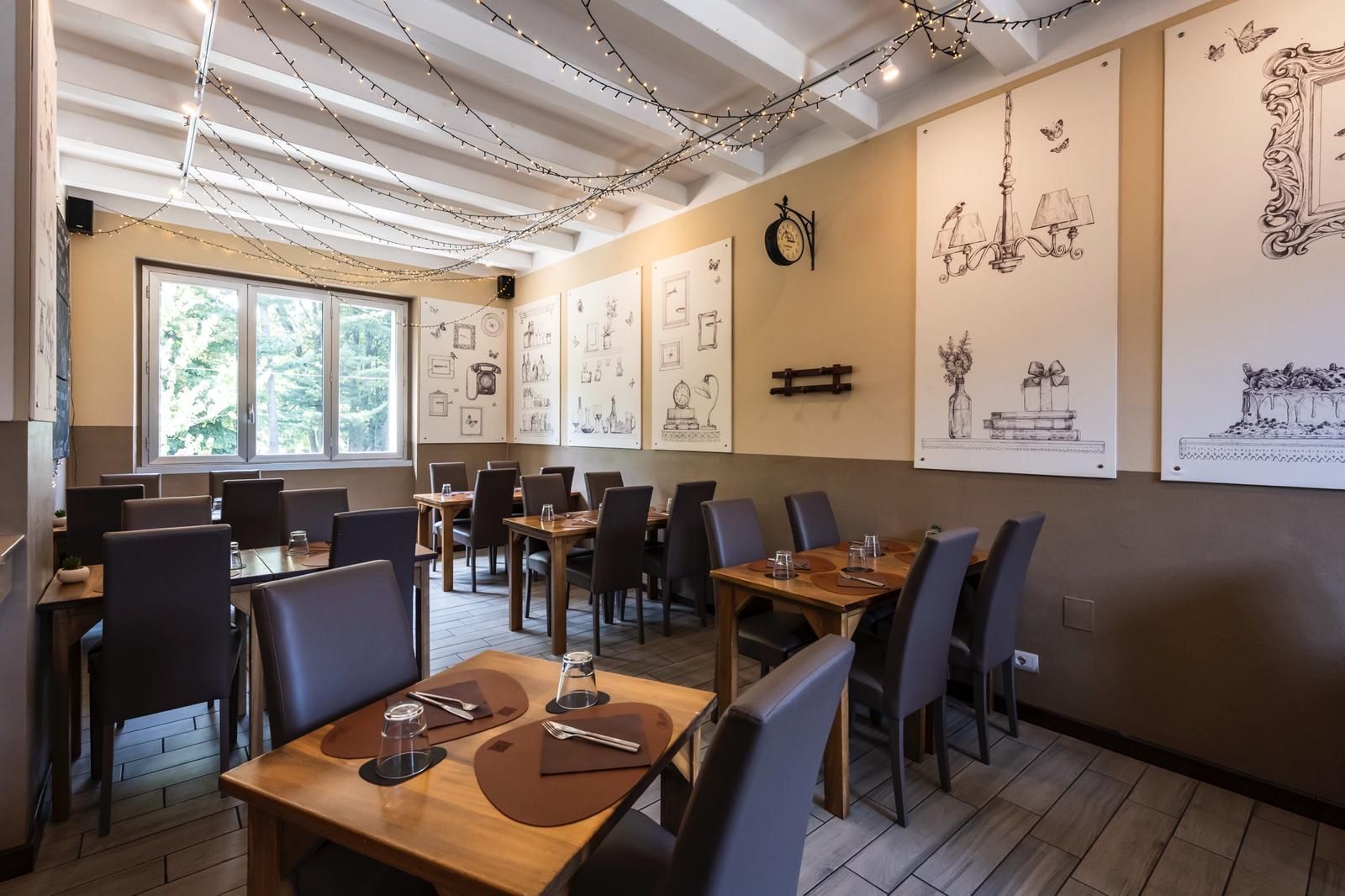 Interno del ristorante: tavoli in legno con apparecchiati, sedie scure, grandi finestre, pareti beige, luci a filo e disegni decorativi.