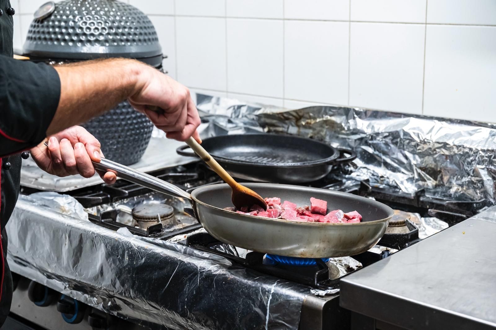 Lo chef mescola la carne in una padella su un fornello a gas; ambiente da cucina.