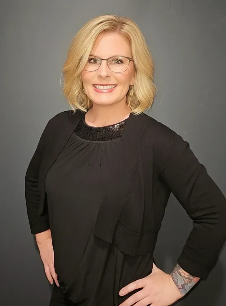 A smiling blonde person with glasses, wearing a black top and cardigan against a gray background.