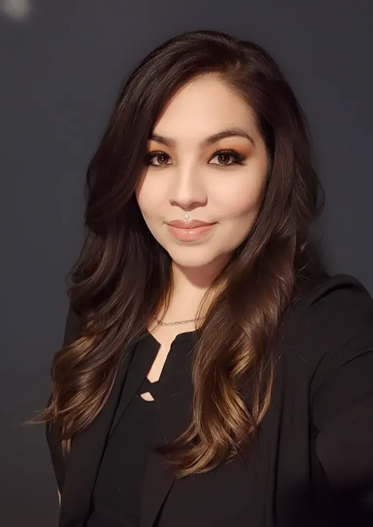 A person with long, wavy dark hair wears a black blazer against a dark, solid-colored background.