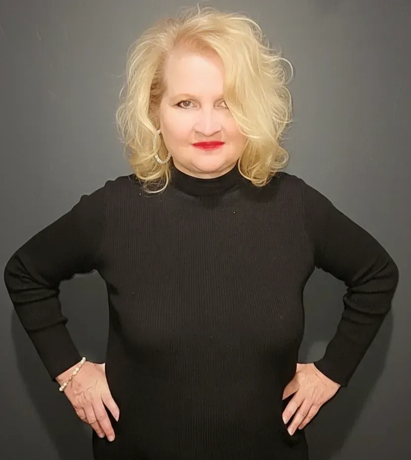 A blonde person with wavy hair wearing a black mock-neck long-sleeve shirt stands against a dark gray background.