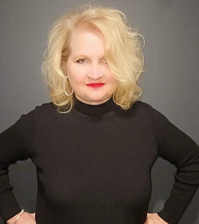 A blonde person with wavy hair wearing a black mock-neck long-sleeve shirt stands against a dark gray background.