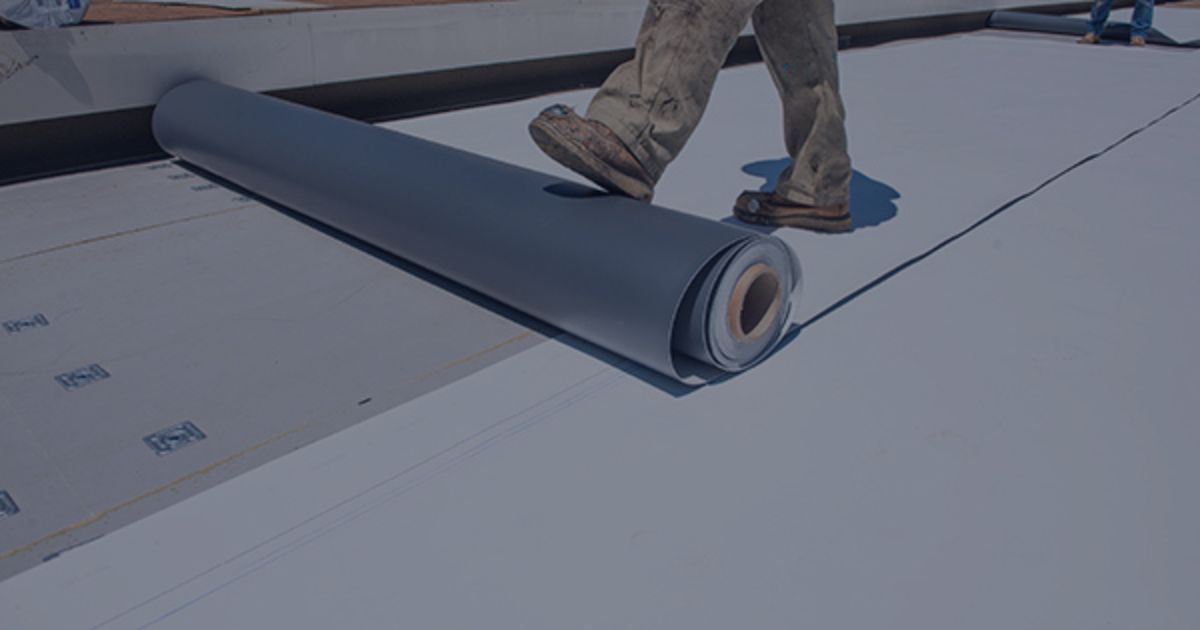 A person in work boots unrolls a large grey waterproof roofing membrane onto a flat roof surface.