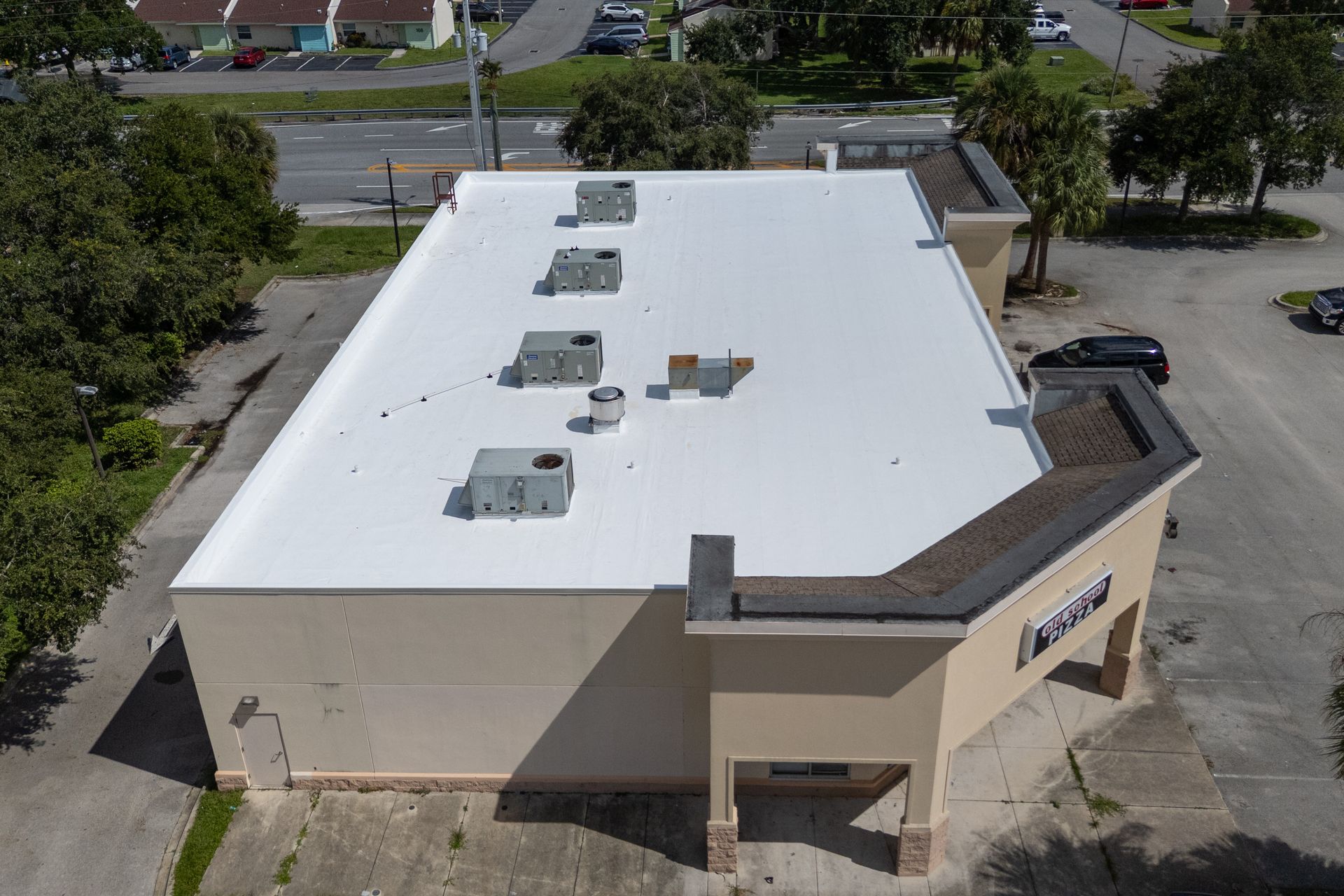 Aerial view of a white community building with a green metal roof surrounded by palm trees, a lawn, and a playground.