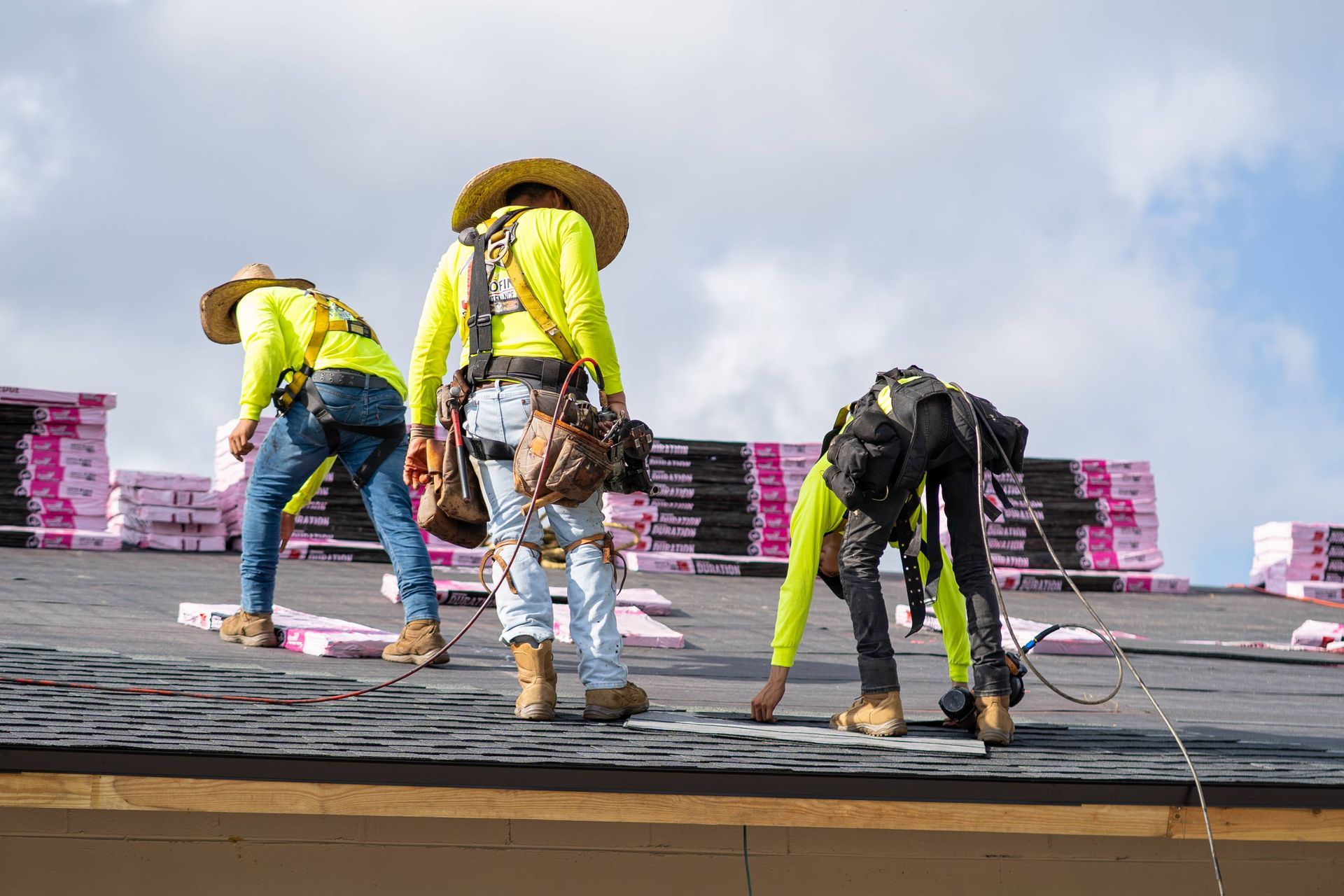 Three roofers in neon yellow shirts and safety harnesses work on a rooftop covered in shingle stacks under a blue sky.