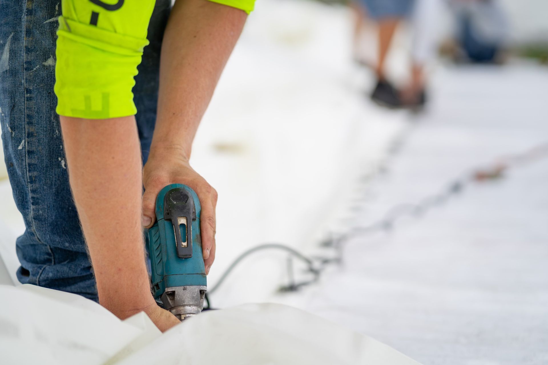 A person in a neon yellow shirt uses a handheld power tool to secure a white tarp to the ground.
