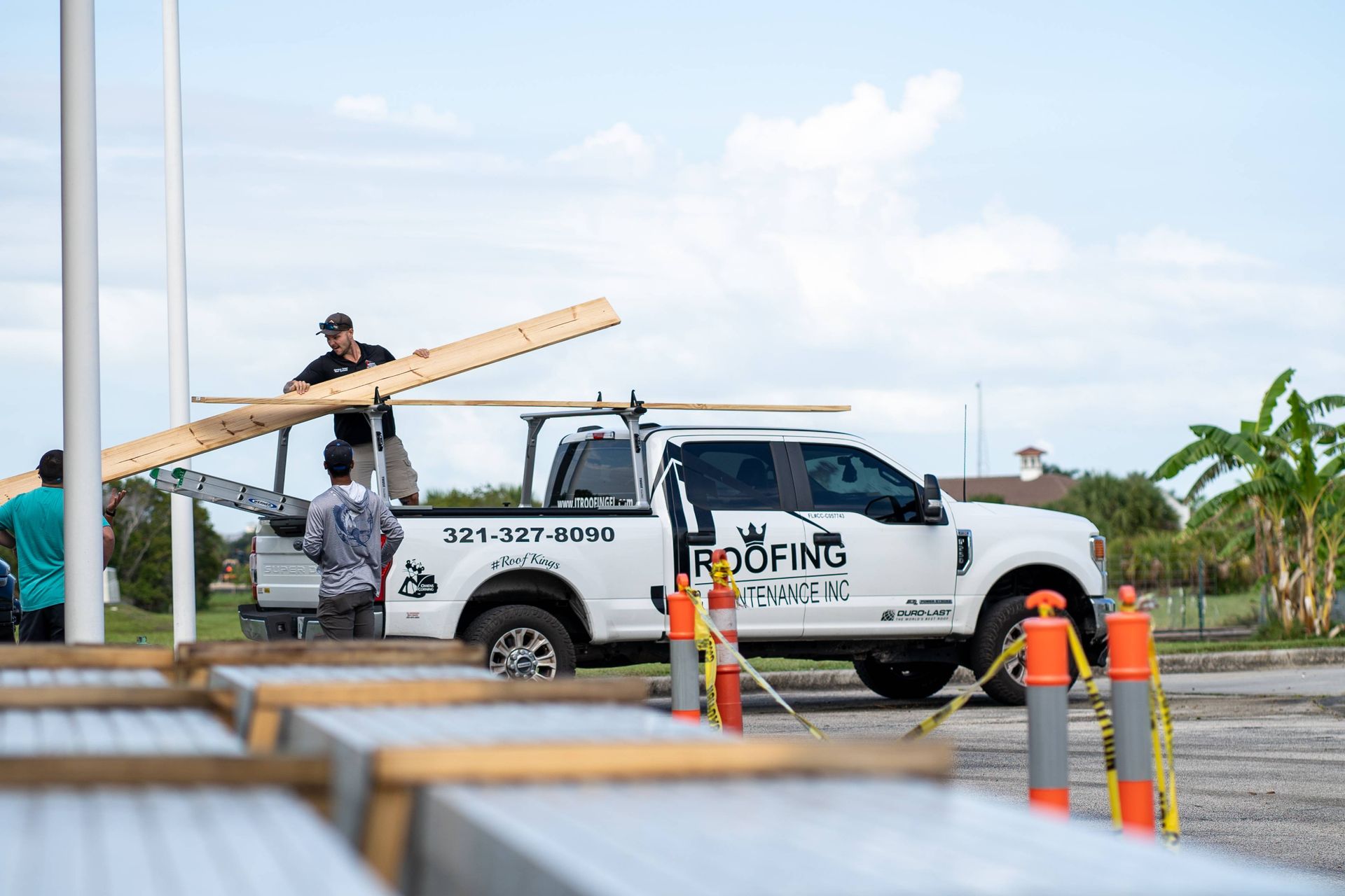 Construction workers load a long wooden plank onto a white roofing company truck parked in an outdoor lot.