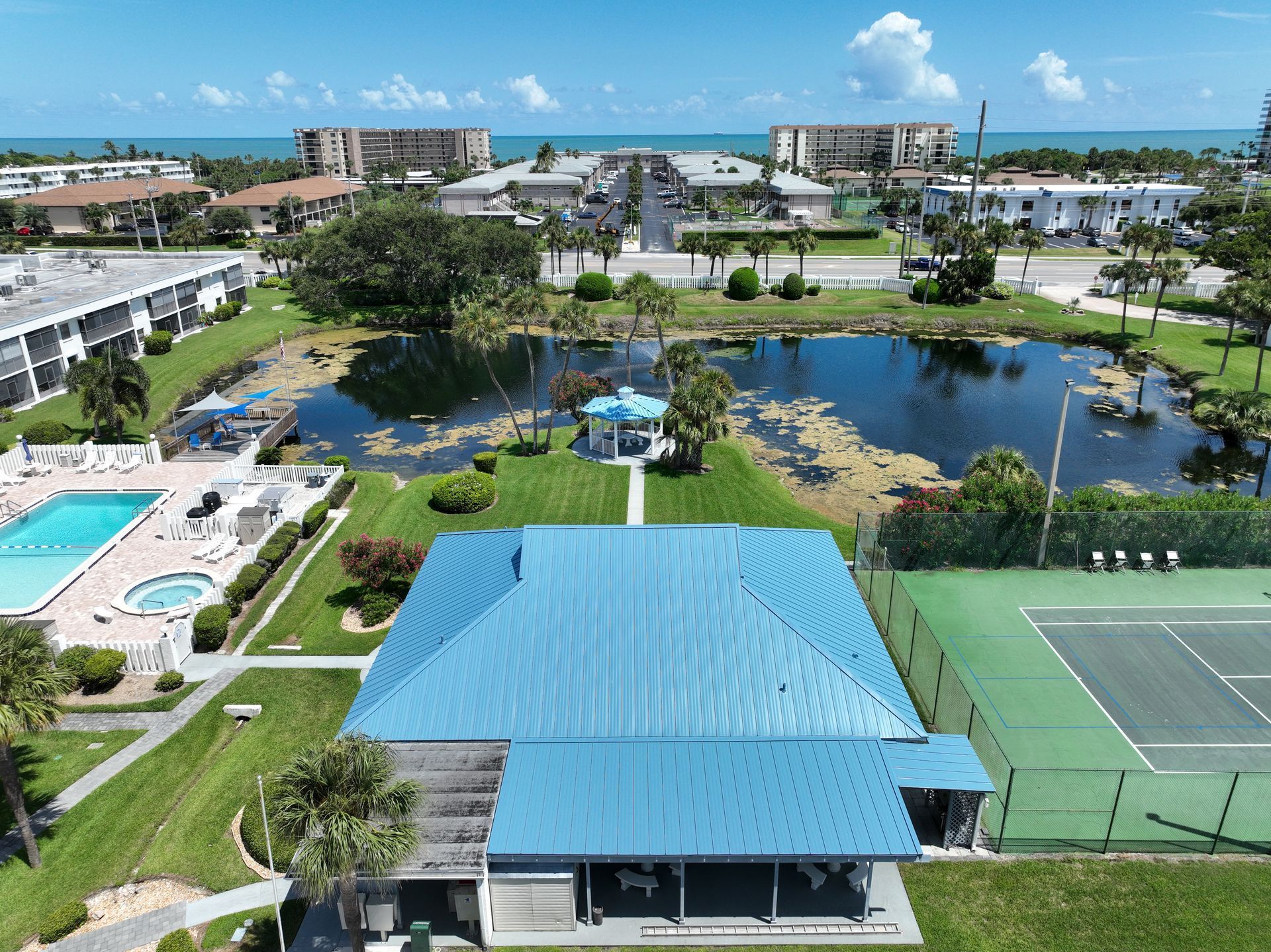Aerial view of a coastal apartment complex featuring a blue-roofed clubhouse, pool, pond, and tennis court.