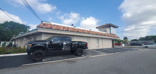 A black pickup truck parked in front of a light-colored commercial building with a tiled roof under a sunny blue sky.