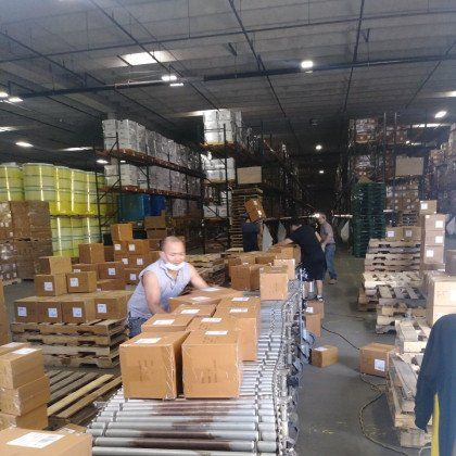 Warehouse workers packing boxes on a conveyor belt, with stacks of boxes and shelves filled with inventory.