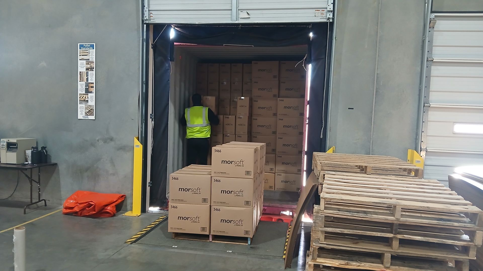 Warehouse loading dock; worker in safety vest loading boxes into a truck. Pallets and boxes are visible.