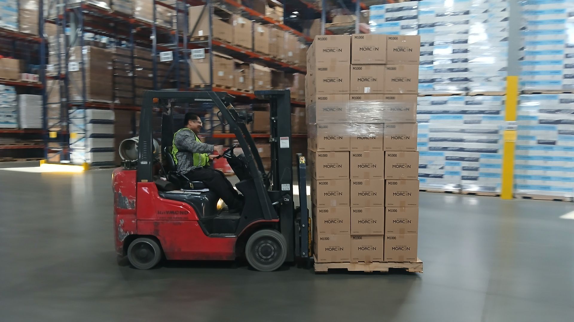 Forklift operator moving a pallet of stacked boxes in a warehouse. Red forklift, tan boxes, man wearing a vest.