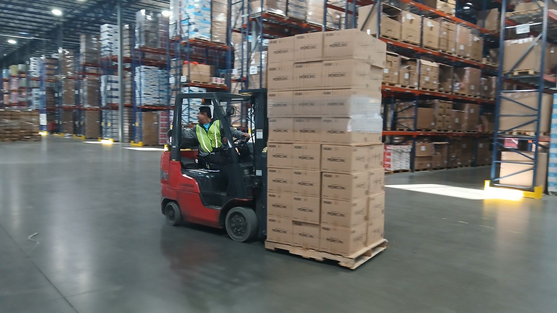 Forklift in warehouse carrying tall stack of cardboard boxes.
