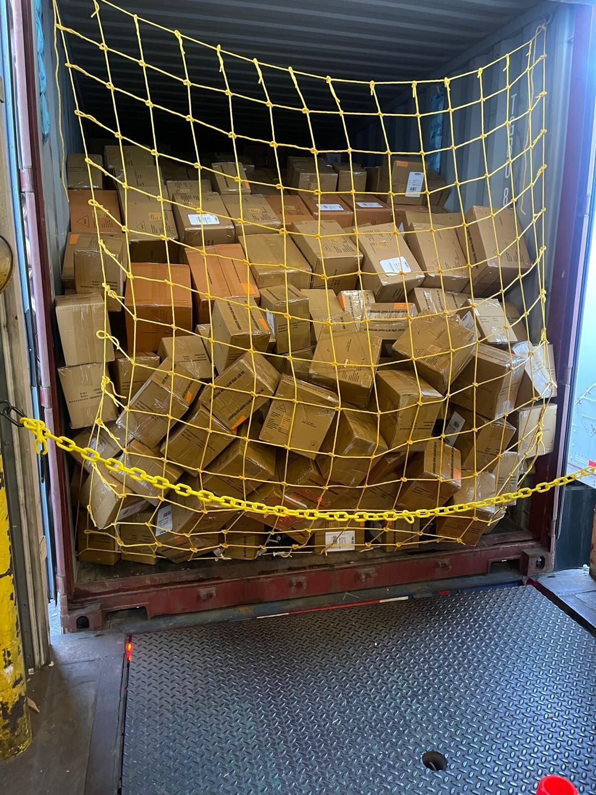 Boxes piled in a shipping container, secured by a yellow cargo net and chain.