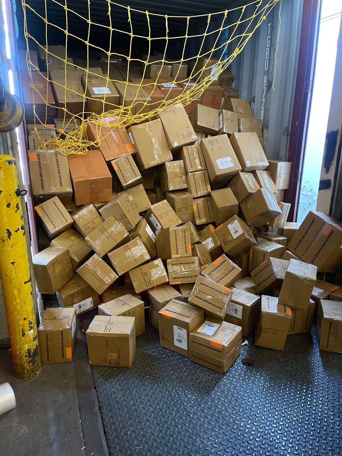 A pile of cardboard boxes tumbled inside a shipping container, secured by a yellow safety net.
