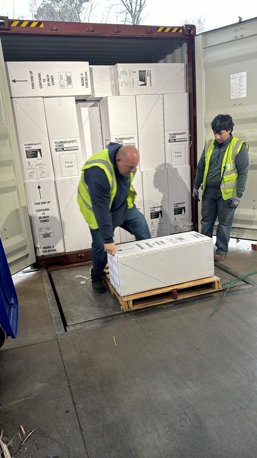 Two men unloading boxes from a shipping container, one man lifts a pallet of boxes.