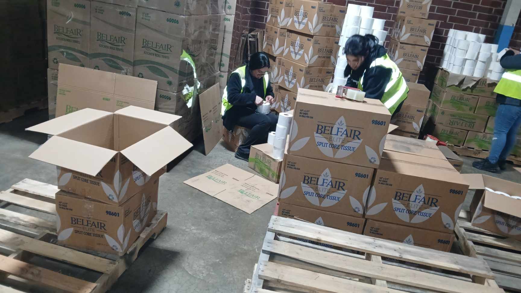 People packing boxes in a warehouse. They wear vests and are surrounded by stacked cardboard boxes and pallets.