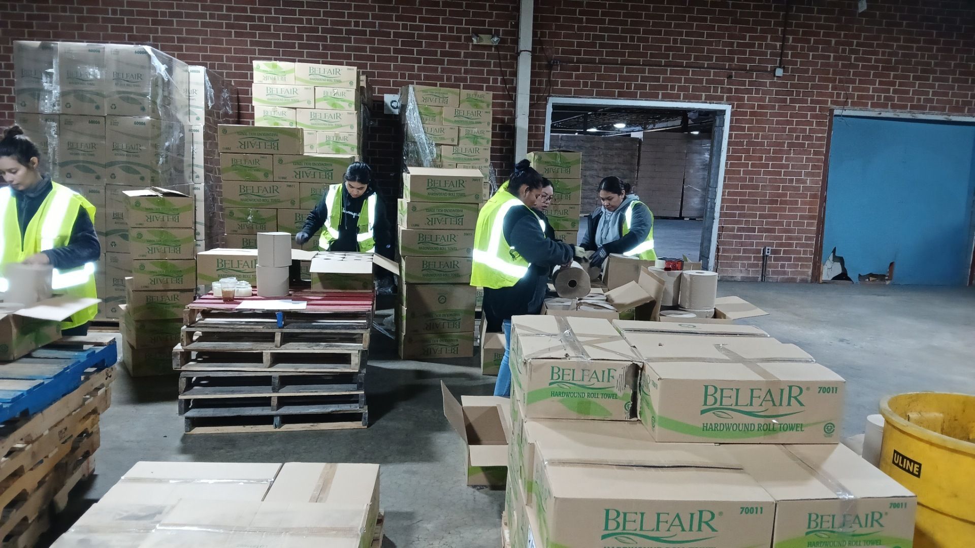 Workers in a warehouse packing boxes, wearing yellow vests, surrounded by stacks of boxes on pallets.